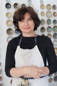 Portrait de Karen Swami devant ses créations de poterie d'art dans son atelier de céramique à Paris, posée devant une fiche où sont collées les exemplaires de ses céramiques d'art en petit format