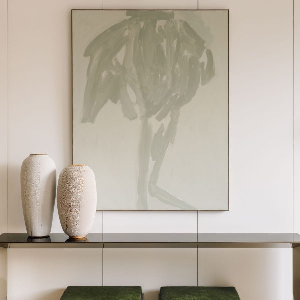 2 tall white ceramic art vases with cracked textures, with gold plated openings and stand, on a reflective metallic glass table, infront of an artistic pastel light green acrylic painting, in a minimalist white room