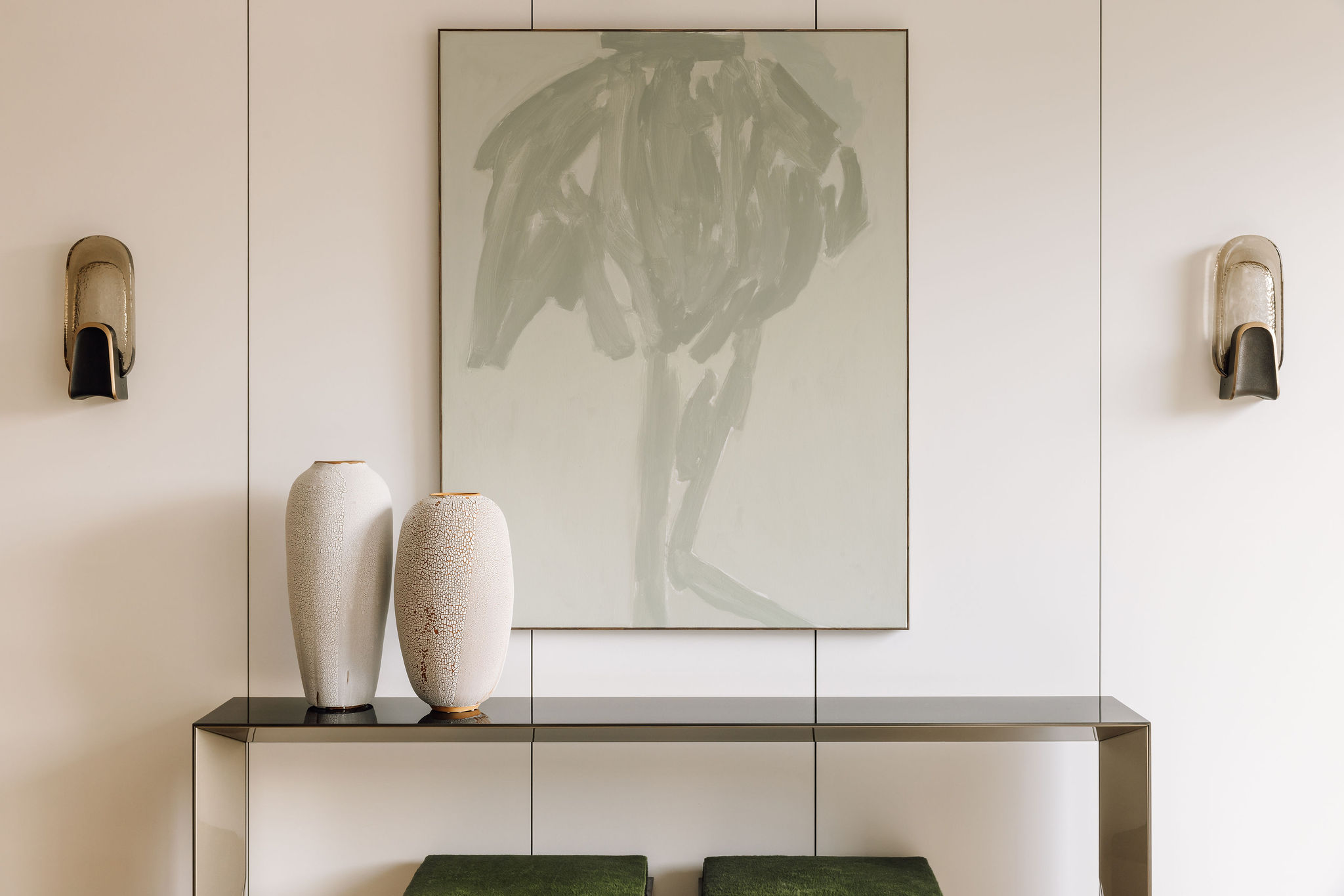2 grands vases d'art en céramique blanche aux textures craquelées, avec des ouvertures et un support plaqués or, sur une table en verre métallique réfléchissant, devant une peinture acrylique vert clair pastel artistique, dans une pièce blanche minimaliste